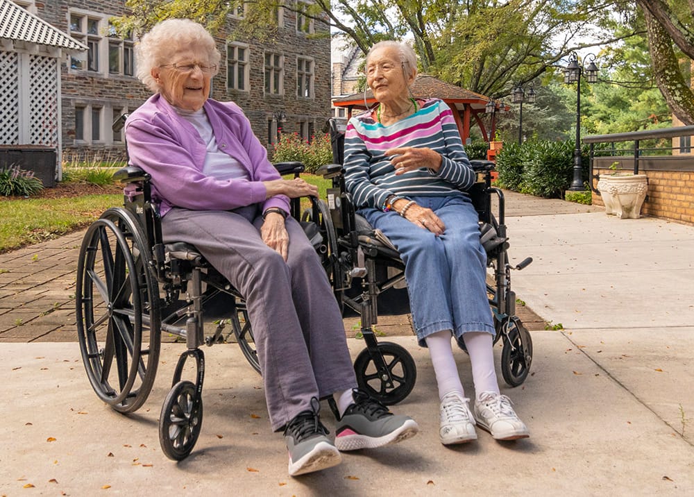 Two elderly women in wheelchairs chatting outside